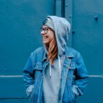 Smiling person in denim jacket and hoodie against a blue wall, exuding casual street style and urban vibe.