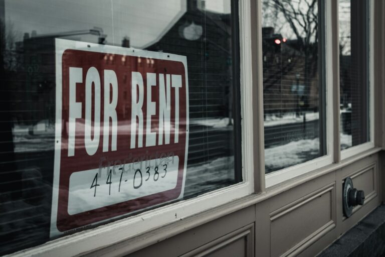 For rent sign in a shop window with winter street reflection, contact number visible for inquiries.