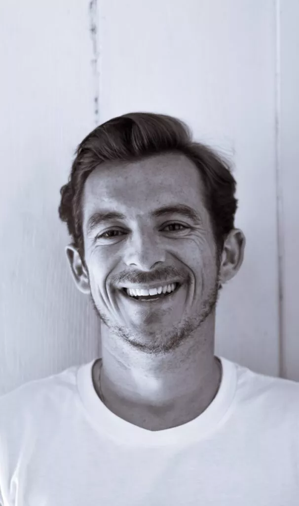 Smiling man in a white shirt against a light wooden background.