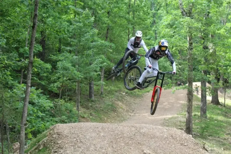 Two mountain bikers performing a jump on a dirt trail surrounded by lush green trees, showcasing extreme sports action.
